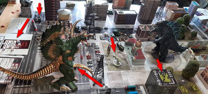Auf einen Blick: Alle Specials, die Godzilla-Tabletop für uns Film-Fans zu bieten hat: Ungeheuer auch in der Mehrzahl, Regelwerk mit Quick-Reference-Bögen (links), Leopard-Panzer, die nicht viel aushalten (oben links), Kleingetier, das für sich genommen bereits die Dimensionen von Ungeheuern erreicht (verglichen mit den Pkw-Modellen), auf dem Hochhausdach rechts unten ein Assault-Team mit schweren Waffen. Die Gittermasten der Überlandleitung sind ebenfalls gut auszumachen. (Foto: Udo Zipelius)